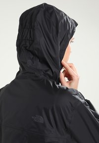 Veste imperméable noire avec capuche, présentant une texture lisse, des détails élastiques sur la capuche et un logo placé à l'arrière.