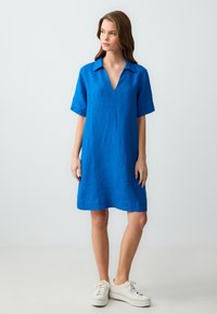 Abito in lino blu con scollatura a V e maniche corte. Presenta una vestibilità rilassata e arriva sopra il ginocchio. Indossato con sneakers bianche.