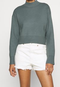 Przycięty sweter w stonowanej zieleni, prążkowany golf i mankiety, miękka tekstura; w połączeniu z białymi szortami dżinsowymi z postrzępionymi brzegami.