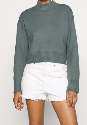 Geknipte trui in gedempt groen, geribbelde col en manchetten, zachte textuur; gecombineerd met witte denim shorts met uitrafeling.