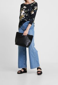 Top a maniche lunghe nera con fantasia floreale e dettagli verdi e rosa, abbinata a jeans a gamba larga blu chiaro e sandali neri flat, mentre tiene una borsa nera.