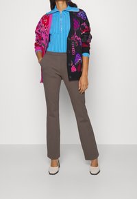 Kolorowy, oversize'owy kardigan z wzorami w różu i czerni, noszony na niebieskim, prążkowanym topie oraz brązowych dopasowanych spodniach, uzupełniony beżowymi butami na blokowanym obcasie.