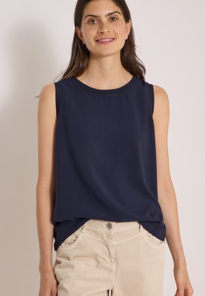 Jeune femme portant un haut sans manches bleu marine et un pantalon beige, souriant doucement, debout devant un fond clair uni.