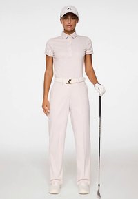 Ljusrosa golfklädsel med en skjorta med krage och skräddarsydda byxor, kompletterad med ett vitt bälte och keps, håller i en golfklubba.