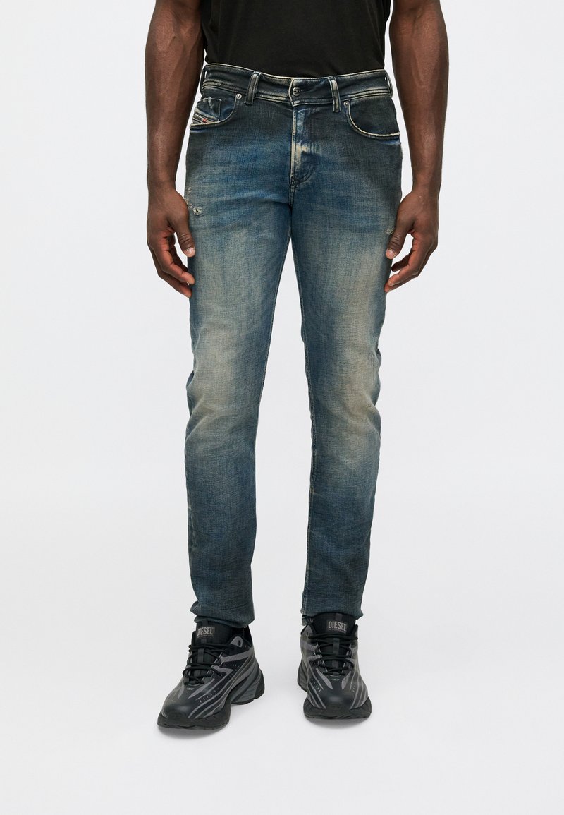 Homem a usar jeans slim-fit azuis desbotados e ténis Diesel pretos, de pé com as mãos relaxadas ao lado do corpo contra um fundo branco simples.