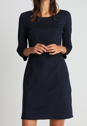 Robe bleu marine en tissu lisse, avec des manches trois-quarts agrémentées de dentelle, un col rond et une silhouette ajustée.
