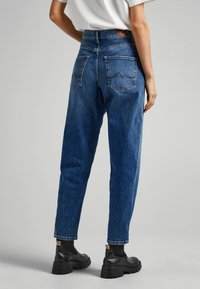 Person som bär högmidjade blå jeans och svarta ankelstövlar, stående mot en enkel ljusgrå bakgrund, sedd bakifrån.