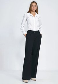 Biała koszula na guziki z ostrym kołnierzykiem, zestawiona z czarnymi spodniami o szerokich nogawkach i wysokim stanie oraz jasnymi szpilkami. Gładka faktura tkaniny.