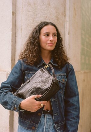 Clutch bag nera con motivo di coccodrillo, dalla forma curvata, texture liscia e zip in hardware dorato. Indossata in modo casual con una giacca di jeans.