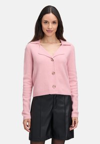 Frau in einem rosafarbenen, geknöpften Cardigan mit weitem Kragen, kombiniert mit schwarzen Leder-Shorts, vor einem einfarbigen weißen Hintergrund.