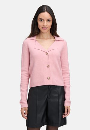 Frau in einem rosafarbenen, geknöpften Cardigan mit weitem Kragen, kombiniert mit schwarzen Leder-Shorts, vor einem einfarbigen weißen Hintergrund.