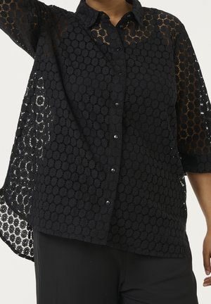 Chemise noire en dentelle à boutons avec des motifs floraux circulaires, portée par une personne levant un bras, montrant le tissu semi-transparent sur les manches et le côté.
