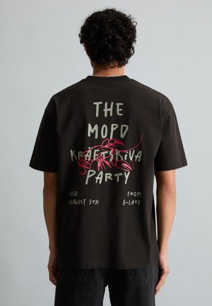 Person mit lockigem Haar, der ein schwarzes T-Shirt mit rotem Hummer und Text trägt, der eine Flusskrebs-Party am 5. August ab 20 Uhr ankündigt.