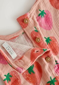 Tela rosa claro con grandes fresas rojas y rosas, botones de madera y una etiqueta de ropa que dice "NEXT Made in India."