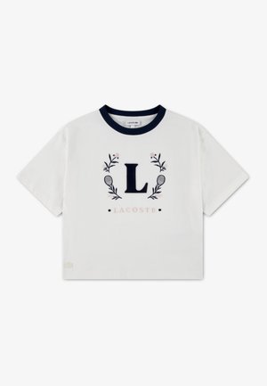 Hvid bomulds-T-shirt med en marineblå rund hals, med et broderet bogstav "L" omgivet af blomsterdetaljer og teksten "LACOSTE" nedenunder.
