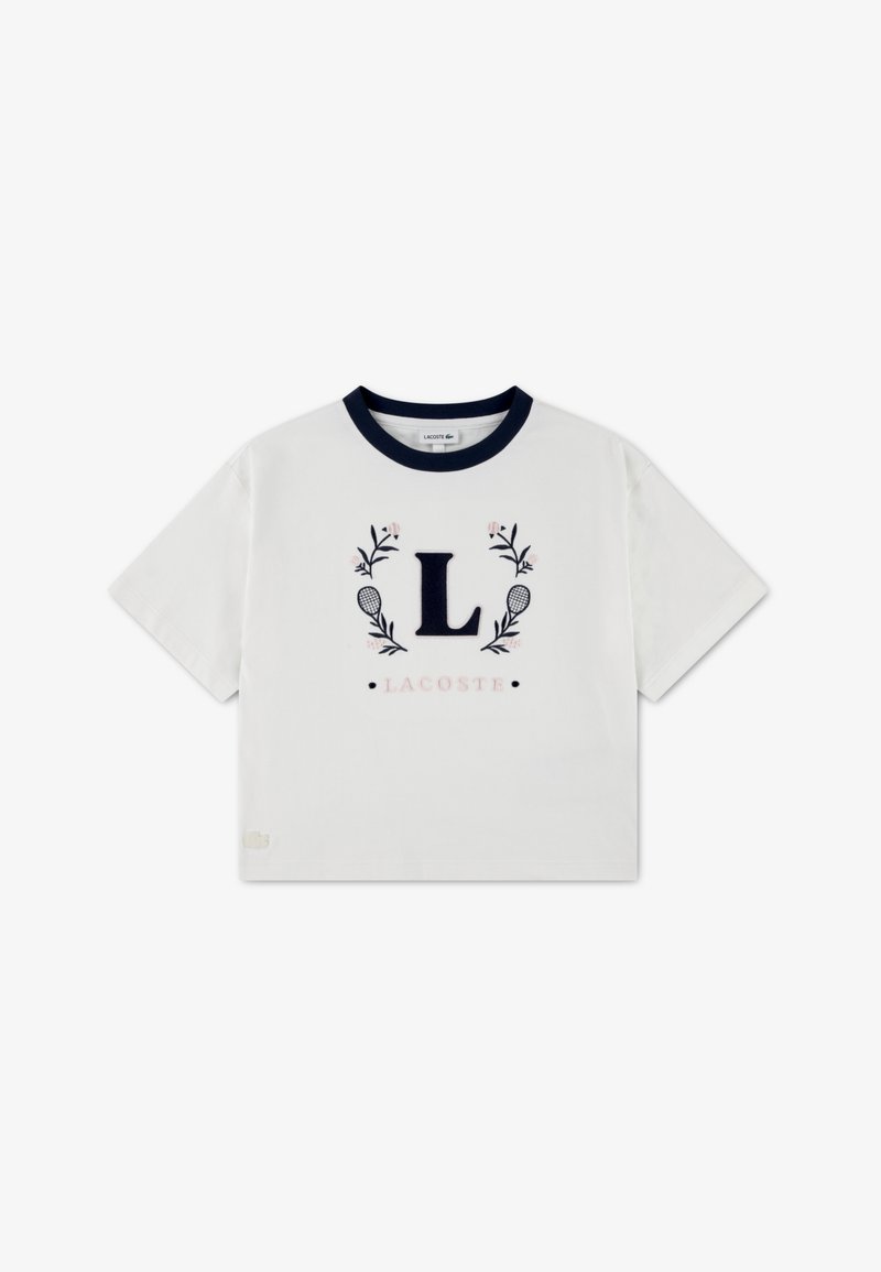 Λευκό βαμβακερό μπλουζάκι με στρογγυλή λαιμόκοψη σε ναυτικό μπλε, με κεντημένο το γράμμα "L" περιβαλλόμενο από ανθισμένα διακοσμητικά και το κείμενο "LACOSTE" από κάτω.