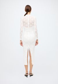 White lace dress featuring a fitted silhouette, long sleeves, and a front slit, with a smooth, solid material below the waist and intricate lace pattern.