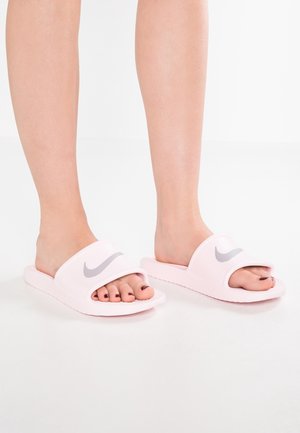 Benen met lichtroze Nike slippers met grijs logo, tenen zichtbaar en donkerrood gelakt, staand op een witte ondergrond.