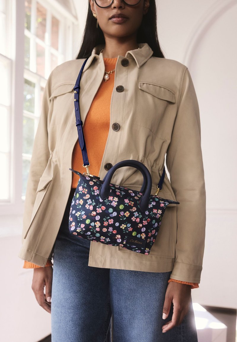 Sac à main floral bleu marine avec un design structuré, en tissu, doté de deux anses et d'une bandoulière amovible, associé à une veste couleur camel et un haut orange.