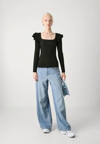 Svart langermet topp med volangdetaljer på skuldrene, kombinert med lyseblå vide jeans og en liten blå håndveske. Hvite joggesko fullfører antrekket.