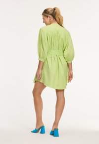 Abito verde lime con maniche a palloncino e cintura in vita; presenta un dettaglio arricciato sulla schiena. Indossato con mules blu con tacco a blocco.