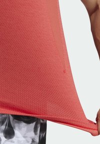 Rød atletisk skjorte med tekstureret stof, der har en sideslids og en løs pasform. Skjorten holdes fra kanten.