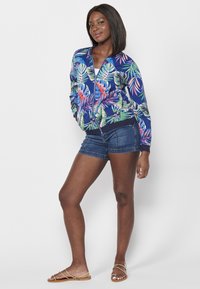 Giacca da bombardiere navy con motivo di foglie tropicali colorate, maniche e orlo a coste. Abbinata a pantaloni shorts in denim e sandali dorati.
