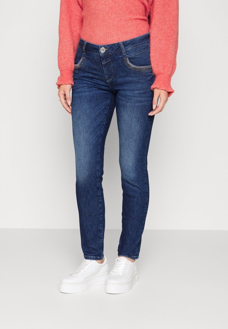 Femme portant un jean slim bleu foncé avec des poches avant cloutées, un pull corail à manches longues et des baskets blanches à plateforme, debout sur un sol blanc.