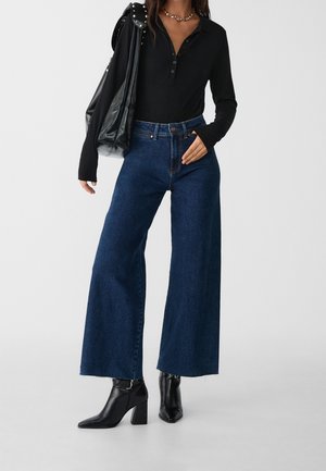 Vrouw draagt een zwarte top met lange mouwen, wijde donkerblauwe jeans, zwarte enkellaarsjes met hak, gouden kettingketting en draagt een zwarte leren tas met studs.