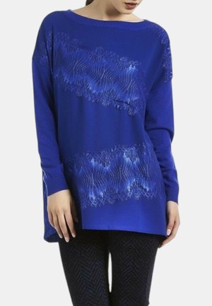 Donna che indossa un maglione lungo blu con bande diagonali di pizzo trasparente, abbinato a pantaloni scuri a fantasia, in piedi davanti a uno sfondo semplice.