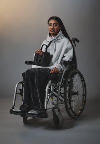 Svart håndveske holdt av en person i rullestol, iført en grå hettegenser med en sløyfedesign og svarte glitrende bukser, sittende mot en nøytral bakgrunn.