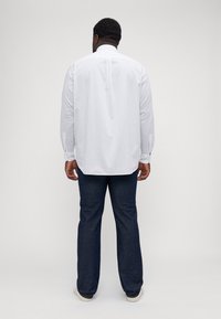 White long-sleeve shirt with a subtle dotted pattern, back pleat detail, paired with dark blue jeans and light-colored sneakers.