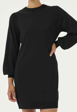 Femme portant une robe noire en maille à manches longues bouffantes, debout avec une main sur la hanche devant un fond uni.