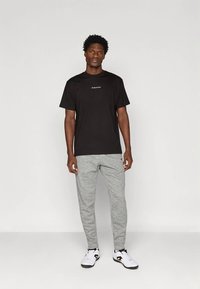 Άνδρας στέκεται μπροστά σε απλή υπόβαθρο, φορώντας μαύρο κοντομάνικο πουκάμισο, γκρι παντελόνια jogger και λευκά αθλητικά παπούτσια.