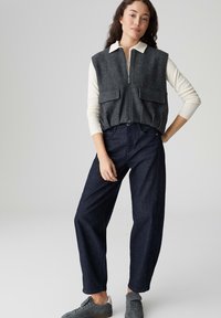 Gilet di lana grigia sopra una camicia crema a maniche lunghe, abbinata a jeans a gamba larga blu scuro e sneakers grigie. Presenta chiusura con zip e tasche frontali.