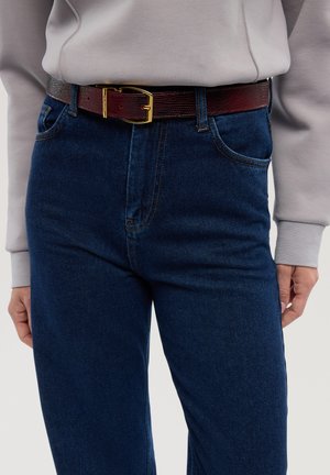 Bruine leren riem met een gestructureerde afwerking en een gouden metalen gesp, gedragen over donkerblauwe spijkerbroek en een lichtgrijze sweatshirt.