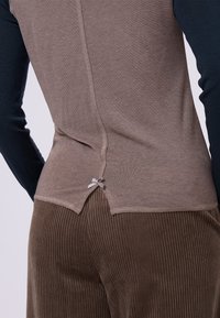 Top marrone chiaro con maniche lunghe blu navy, dotato di una chiusura posteriore con un piccolo fiocco, abbinato a pantaloni marroni a costine. Tessuto morbido e textured.