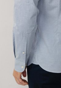 Svetlomodrá károvaná košeľa s dlhým rukávom, lakťovými záplatami a zapínaním na gombíky, ktorú nosí osoba s tmavými nohavicami na neutrálnom pozadí.