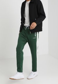 Gröna träningsbyxor med vita sidostripes, dragsko i midjan, tillsammans med en svart jacka och vita sneakers, med en avslappnad passform.