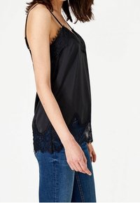 Svart satäng camisole med spetsdetaljer, som har tunna band, en scalloped nederkant och en flytande silhuett. Matchas med blå denimjeans.