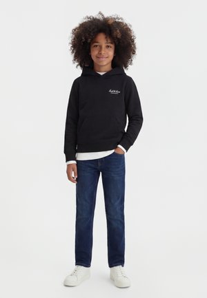 Enfant aux cheveux bouclés portant un sweat à capuche noir, un jean bleu et des baskets blanches, debout avec une main dans la poche sur fond blanc.