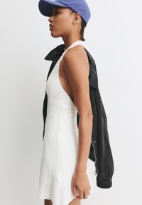 Robe blanche texturée à design sans manches et décolleté. Veste en velours côtelé noire portée sur l'épaule. Casquette bleue portée légèrement en arrière.