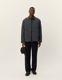 Grijze button-up jas met een gestructureerde afwerking en een enkele borstzak, gecombineerd met donkere broeken en een geblokte pet.