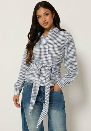 Femme portant une chemise à carreaux blanche et bleue à manches longues nouée à la taille et un jean bleu avec des poches, debout devant un fond uni.