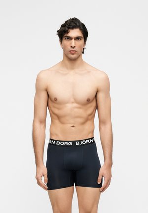 Homme musclé debout, portant un boxer noir de la marque Björn Borg sur un fond blanc uni.