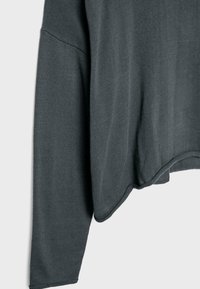Pull en tricot gris foncé à manches longues avec couture d'épaule descendue et ourlet légèrement arrondi sur un fond clair uni.