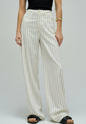 Mujer con pantalones blancos de talle alto con finas rayas negras verticales, sandalias negras, top negro y pulsera dorada en la muñeca derecha.