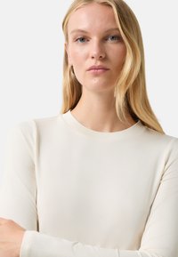Langärmliges Oberteil mit Rundhalsausschnitt aus weichem, leichtem cremefarbenen Stoff. Glatte Textur mit minimalistischem Design und ohne sichtbare Muster oder Akzente.