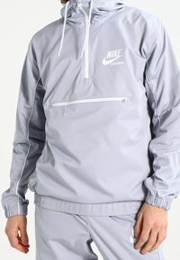 Light grey Nike windbreaker featuring a half-zip design, adjustable hood, and front pocket with a white logo and contrasting seams.