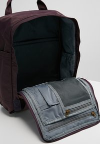 Backpack in deep burgundy fabric with a textured finish. Interior features grey compartments with mesh pockets and zip closures.